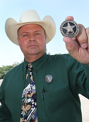 A man in a dress shirt with a light colored cowboy hat one holding up a silver and black circle star badge