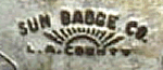 A close up of the back of a silver badge with the words "Sun Badge Co." above an image of the rising or setting sun with the words "L.A. County" underneath it.