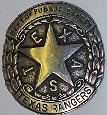 A silver circular badge with a star in the center and leaves along the side. Alternating with the points of the star are the letters that spell "Texas" across the top of the star are the words "Dept. of Public Safety". Across the bottom of the circle are the words "Texas Rangers".