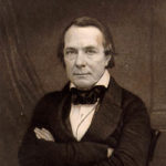 A black and white waist up photograph of a clean shaven man in a dark suit with his arms crossed.