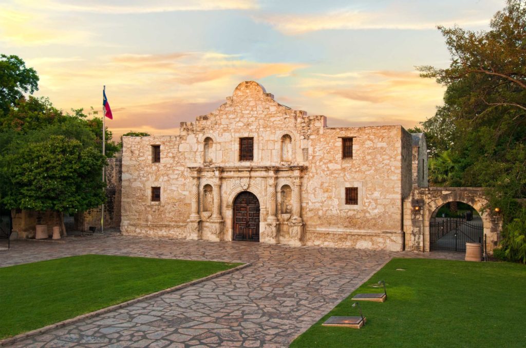 A stone building in the Spanish mission style with a large stone and grass courtyard flying a Texas flag against the setting or rising sun.