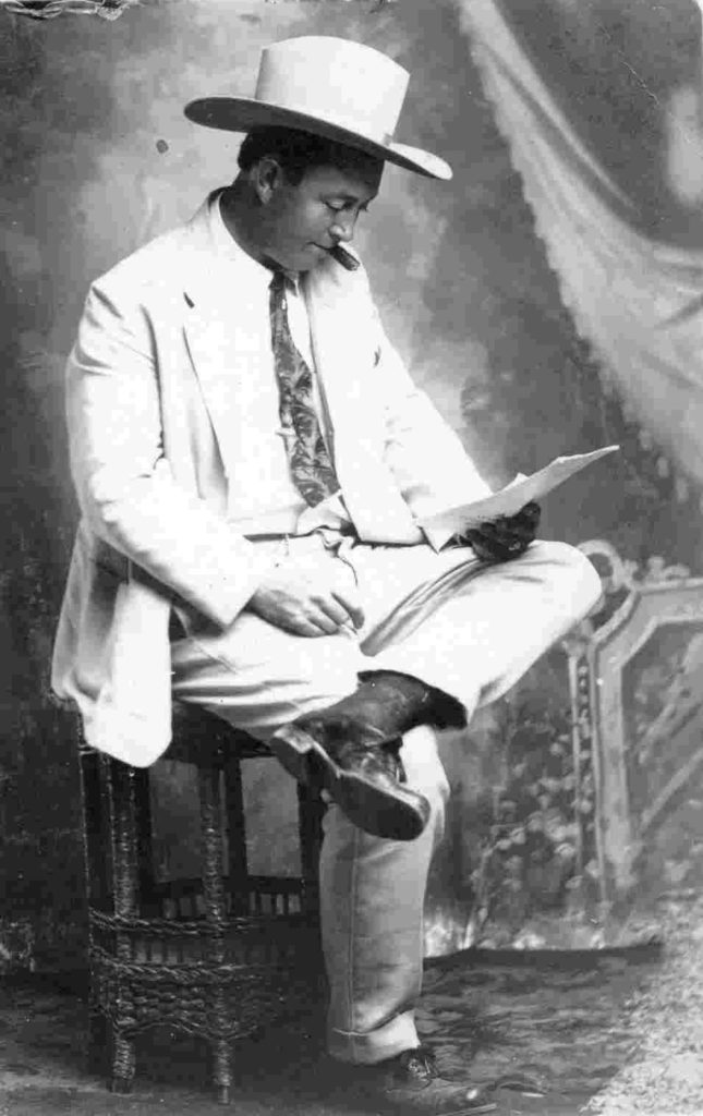 A man in a white cowboy hat and a light colored suit sitting on a stool, reading a paper, and smoking a cigar.