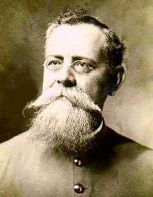 A black and white head shot of a man with glasses a beard and a large mustache.