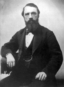 A waist up picture of a man in a dark suit with a beard and a pinkie ring.