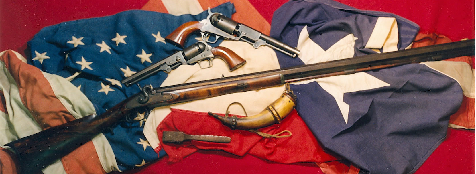 Two pistols, a rifle, a powder horn, and another tool arranged across an American and Texas flag.