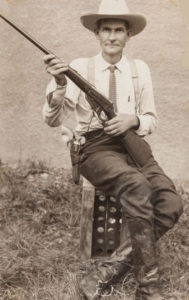 A 1900s Ranger in a collared shirt, tie, cowboy boots, and a cowboy hat poses for a picture with his rifle.