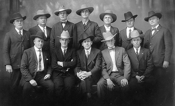 Twelve men posing for a picture in 1920s dark suits and cowboy hats.