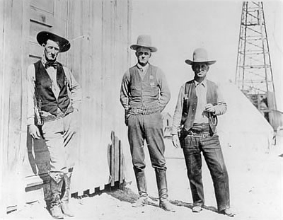 Three early 1900s Rangers in vests and cowboy hats standing in front of a building with an oil rig in the background.