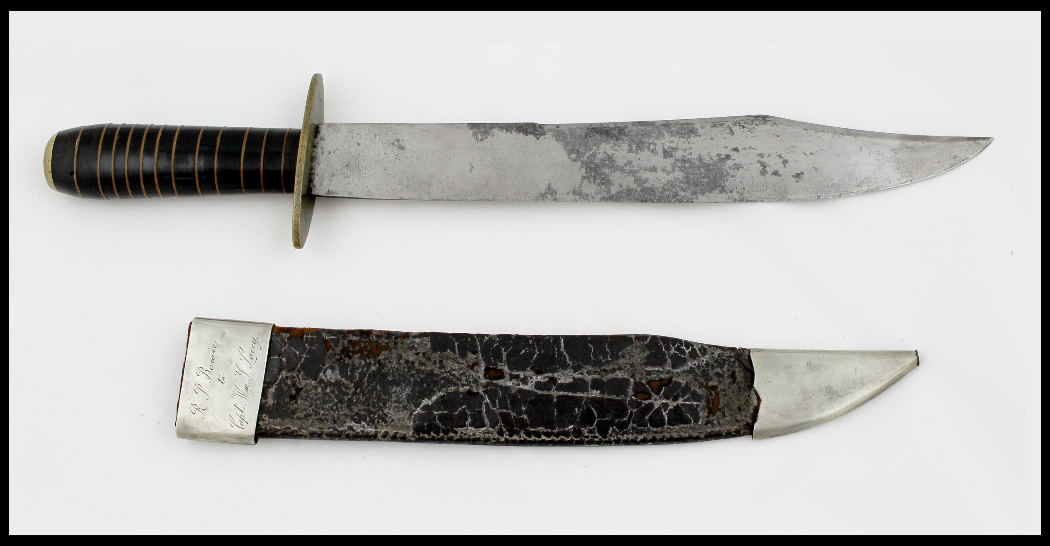 A silver metal bowie knife with a black and bronze colored handle above a silver metal and black leather scabbard with engraving on the top.