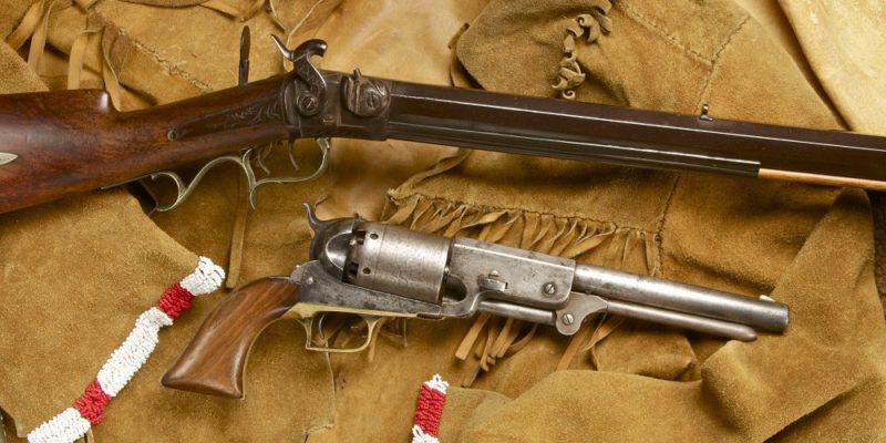 A rifle with an intricate brass trigger guard and pistol with wooden grips arranged across a fringed and beaded suede jacket.