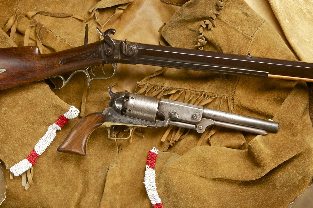 A rifle with an intricate brass trigger guard and pistol with wooden grips arranged across a fringed and beaded suede jacket.