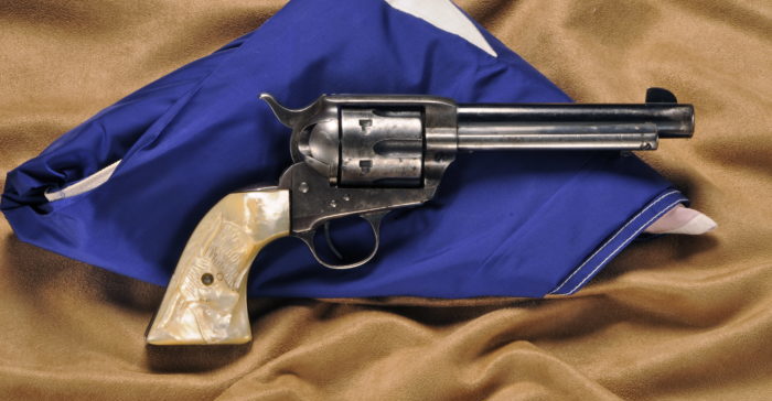 A silver metal pistol with mother of pearl grips engraved with a long horn. The firearm is resting against a folded up Texas flag.