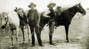 Two armed men in 1800s Ranger garb complete with hats standing next to their horses.