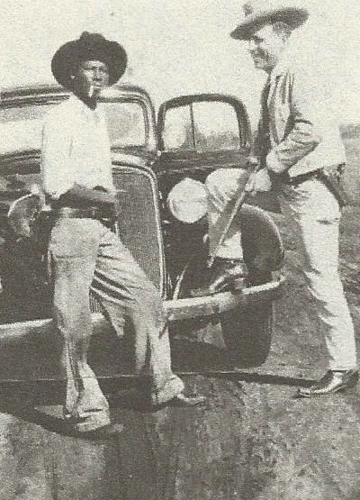 Two men standing in front of a vintage car. One man is holding a firearm.