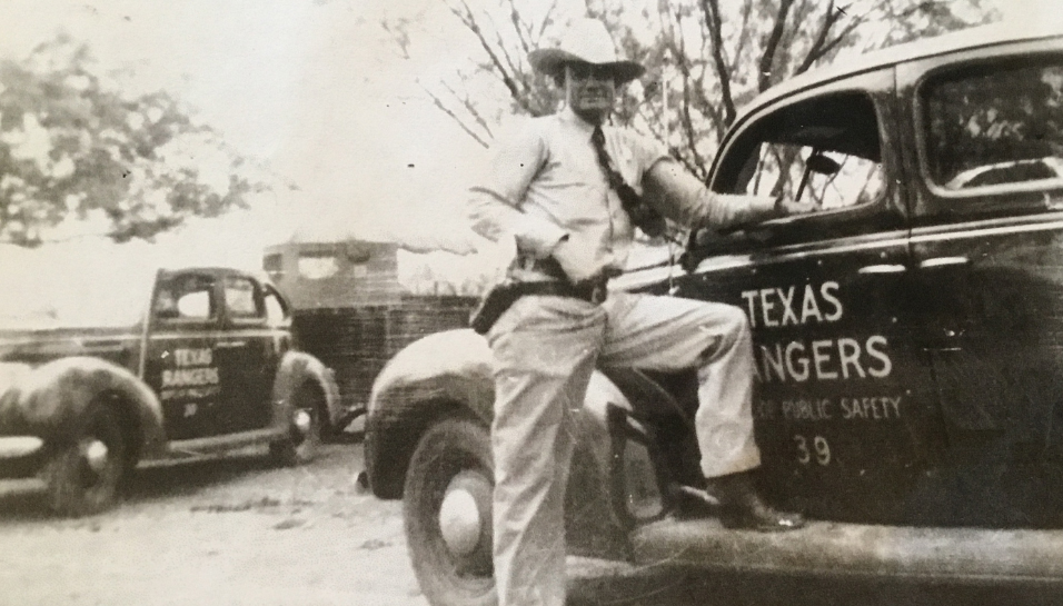 A man in a white hat, light colored pants and button up shirt with tie, and a gun belt and cowboy boots standing next to a car with the words "Texas Rangers Dept. of Public Safety 39" on it.