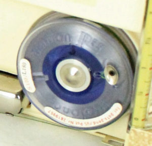 A grey and blue circle covered in clear plastic attached to a cream tan background with a ruler.