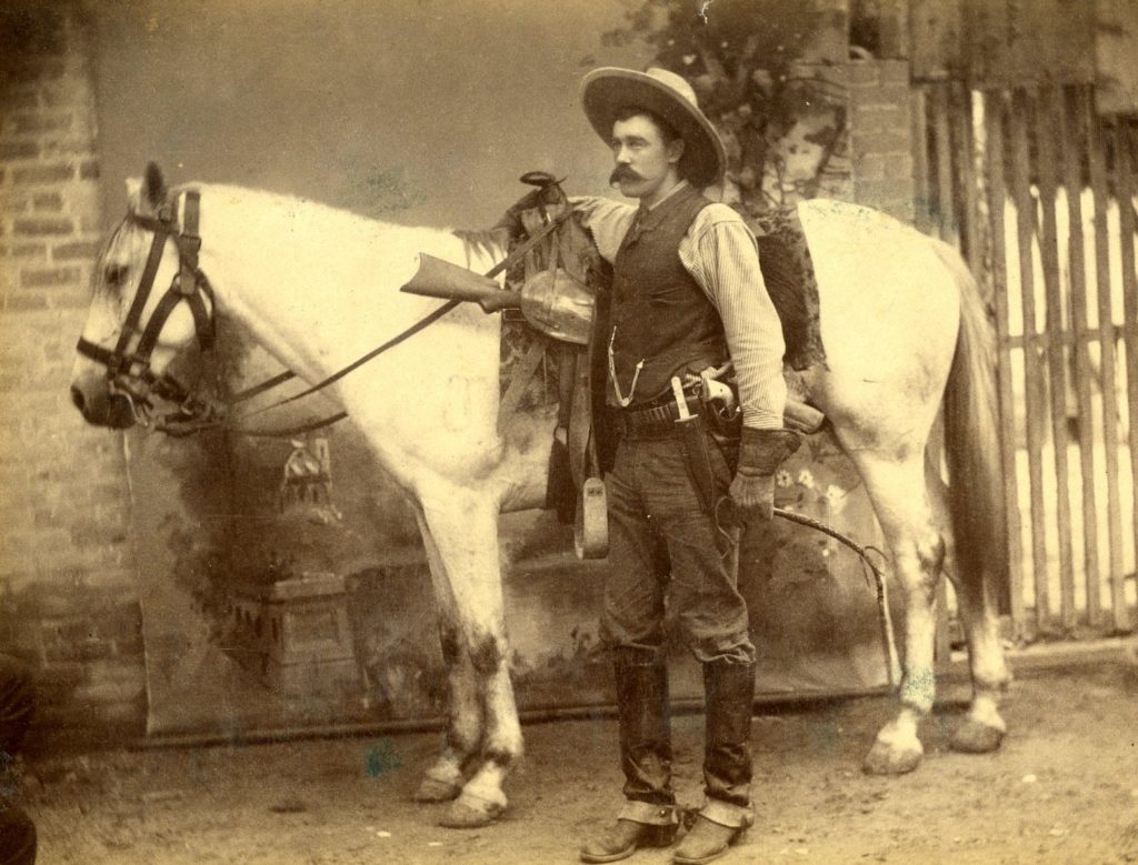 A man with a large mustache and cowboy hat dressed in 1800s Ranger gear standing next to a light colored horse.