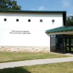 A white building with a green roof and a tan stone rim around the bottom. Written on the building are the words "Texas Rangers Education Center".